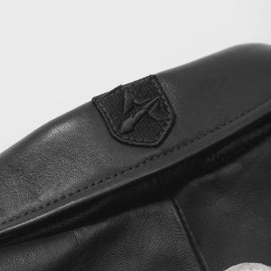 Pu&ograve; includere: Primo piano di un articolo in pelle nera con una cucitura. Una piccola toppa ricamata con un emblema stilizzato &egrave; cucita sulla pelle. La pelle ha una consistenza e una lucentezza sottili, che suggeriscono un materiale di alta qualit&agrave;.