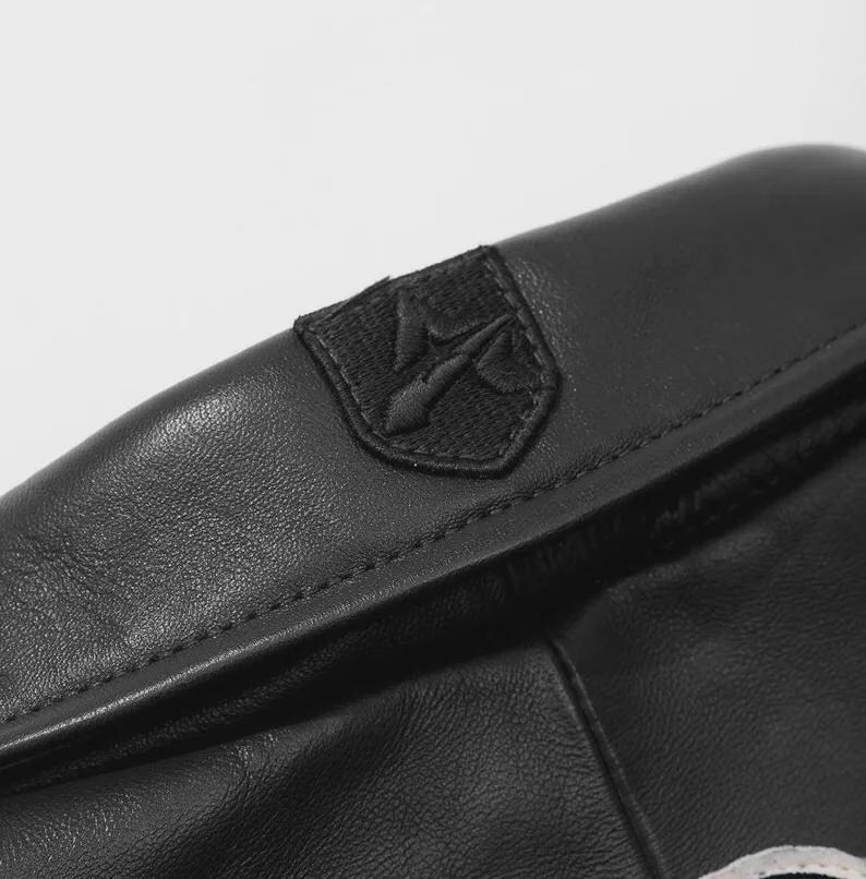 Pu&ograve; includere: Primo piano di un articolo in pelle nera con una cucitura. Una piccola toppa ricamata con un emblema stilizzato &egrave; cucita sulla pelle. La pelle ha una consistenza e una lucentezza sottili, che suggeriscono un materiale di alta qualit&agrave;.