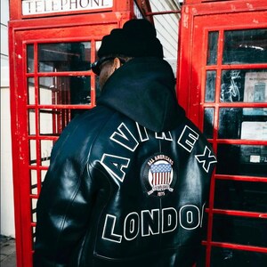 Puede incluir: Chaqueta de cuero negra con letras blancas que deletrean "AVIREX" y "LONDON" en la espalda. Un parche con un diseño de bandera estadounidense está centrado encima de "LONDON". La chaqueta se usa frente a dos cabinas telefónicas rojas.