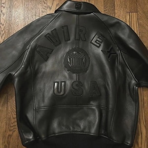 May include: Black leather bomber jacket with the embossed text "AVIREX USA" and a circular emblem on the back. The jacket has a ribbed hem and cuffs, and a classic collar. The jacket is displayed on a wooden surface.