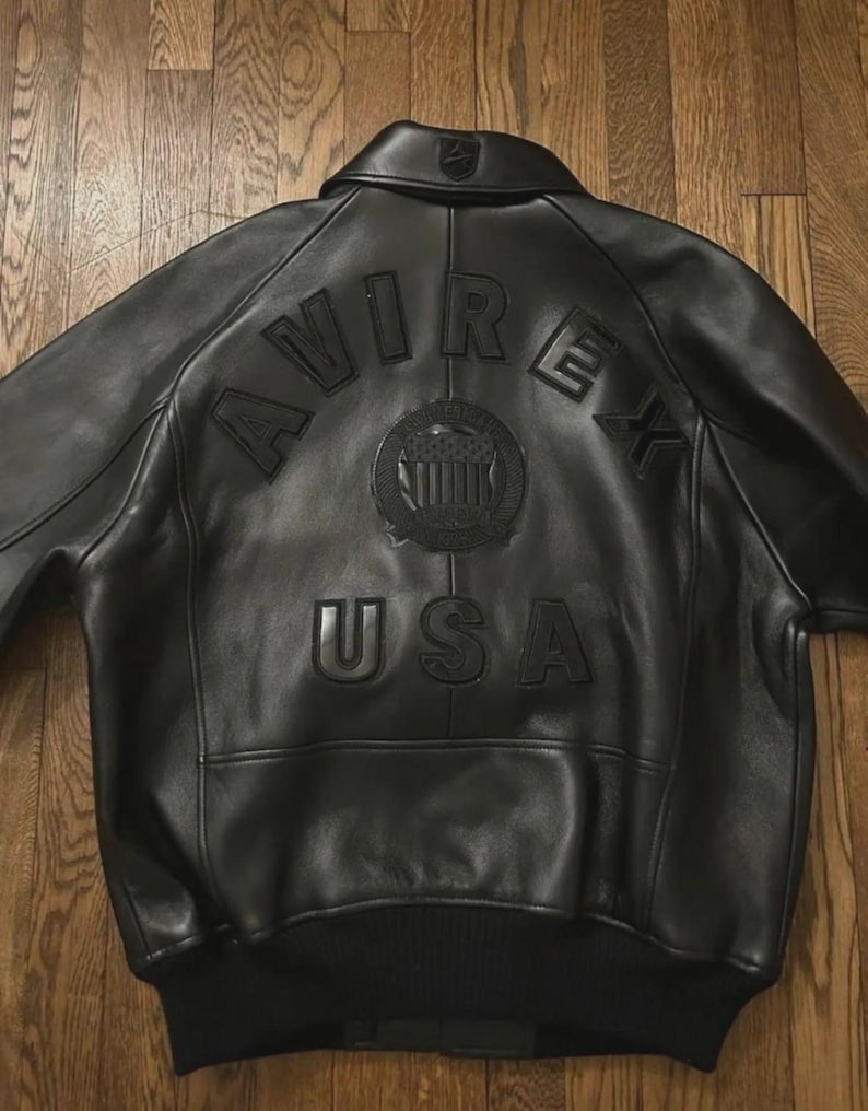 May include: Black leather bomber jacket with the embossed text "AVIREX USA" and a circular emblem on the back. The jacket has a ribbed hem and cuffs, and a classic collar. The jacket is displayed on a wooden surface.