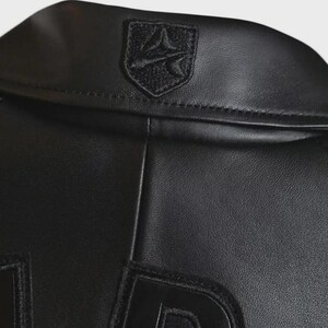 May include: Close-up of a black leather jacket. The jacket features a small, embroidered emblem on the collar and large, raised lettering on the lower portion. The leather has a subtle texture, and the stitching is visible.
