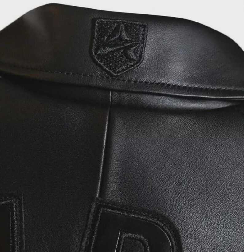 May include: Close-up of a black leather jacket. The jacket features a small, embroidered emblem on the collar and large, raised lettering on the lower portion. The leather has a subtle texture, and the stitching is visible.