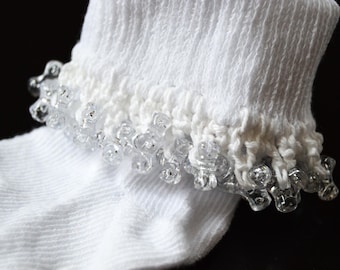 May include: Close-up of a white sock with a decorative, clear, beaded trim around the cuff. The sock is made of a ribbed knit fabric. The trim is made of clear, droplet-shaped beads.