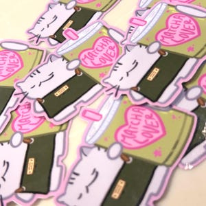 Op de afbeelding: Een verzameling gestanste stickers met een witte kat met grijze strepen die een groen matchadrankje in een doorzichtige beker vasthoudt. De beker heeft een roze hartje met de tekst MATCHA LOVER. De kat heeft een kalme uitdrukking.