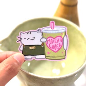 Op de afbeelding: Een gestanste sticker met een witte kat met grijze strepen die een donkergroen schort draagt en een grote groene matchadrank in een plastic beker met een roze rietje vasthoudt. De beker heeft een roze hartje met de tekst MATCHA LOVER erop.