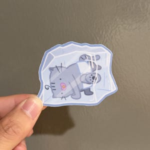 May include: A die-cut sticker featuring a light gray cartoon cat with a small pink nose and dark gray stripes, depicted as being trapped inside a translucent, light blue block of ice with visible geometric facets and a light blue border.
