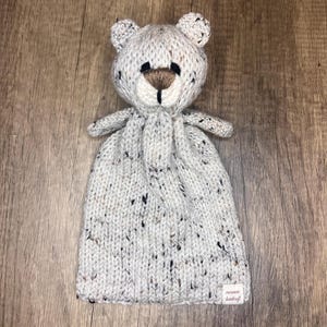 Può includere: Un cappello da bambino a forma di orso, lavorato a mano, grigio chiaro, con macchie nere e marroni. L'orso ha un naso marrone e occhi neri. Il cappello ha piccole orecchie e braccia. Una piccola etichetta con testo è cucita in basso.