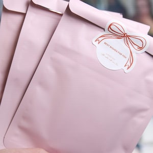 May include: Three light pink gift bags with a rolled top and a sticker that says "Best Wishes For You" with a red bow. The bags are made of a shiny material.
