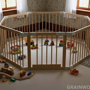 May include: A wooden octagonal playpen with vertical bars, filled with colorful toys. A wooden bookshelf with books and stuffed animals is to the left. The playpen is on a light-colored floor, with a rug and armchair nearby.