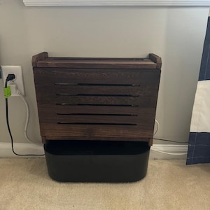May include: A dark brown wooden storage bin with a black plastic base. The bin has a slatted front design and a hinged lid. The bin is sitting on a beige carpet, near a wall with a white trim and an electrical outlet.