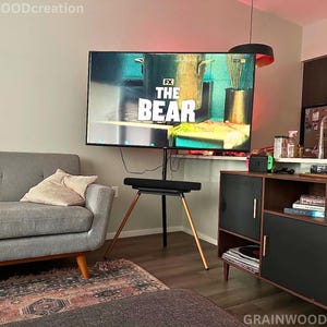 May include: A living room setup featuring a large television displaying "THE BEAR" on the screen. The TV is mounted on a black stand with wooden legs. A gray sofa, a patterned rug, and a dark wood cabinet with gold handles complete the scene.