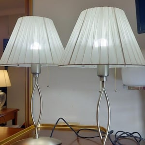 Puede incluir: Dos lámparas de diseño vintage de los años 90 con pantallas plisadas blancas y bases plateadas. Las lámparas tienen un diseño de tallo curvo único y están colocadas sobre una superficie de madera. El texto "Pujol Iluminacióo Barcelona" es visible.