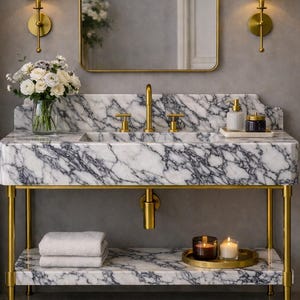Puede incluir: Un mueble de baño con encimera y estante de mármol blanco con vetas gris oscuro, sostenido por un marco de metal dorado. Incluye un grifo dorado, un espejo rectangular, dos apliques de pared, toallas blancas, velas y un florero con flores.