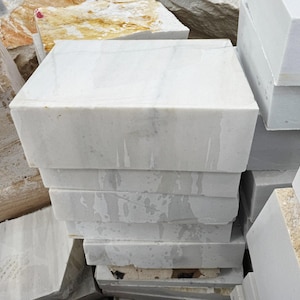 May include: A stack of rectangular white marble blocks. The blocks have a smooth, polished surface with subtle gray veining. The edges are sharp and clean, and the blocks are stacked neatly. Other stone blocks are visible in the background.