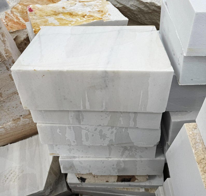 May include: A stack of rectangular white marble blocks. The blocks have a smooth, polished surface with subtle gray veining. The edges are sharp and clean, and the blocks are stacked neatly. Other stone blocks are visible in the background.