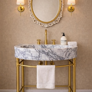Puede incluir: Un lavabo de mármol con grifos dorados y un soporte dorado a juego. Una toalla blanca cuelga debajo del lavabo. Arriba, un espejo ovalado con marco dorado y dos apliques con pantallas plisadas.