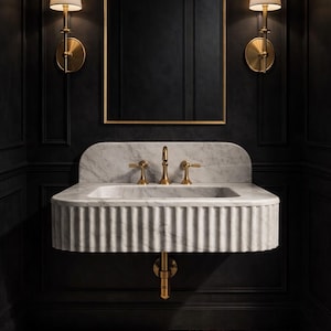 May include: A luxurious bathroom interior featuring a marble sink with gold fixtures, set against a black paneled wall. Two sconces with cream-colored shades flank a gold-framed mirror. The sink has a ribbed design.