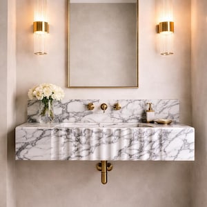 May include: A stylish bathroom interior featuring a marble vanity unit with a scalloped edge and gold fixtures. A gold-framed mirror hangs above the sink, and two gold and glass wall lights are positioned on either side. A vase of white roses sits on the marble countertop.
