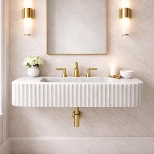 Op de afbeelding: Een moderne badkamer met een witte, gefreesde marmeren wastafel en gouden armaturen. De wastafel is aan de muur bevestigd, met een bijpassende gouden kraan en afvoerbuis. Een goudomrand spiegel hangt boven de wastafel, geflankeerd door twee gouden wandkandelaars.