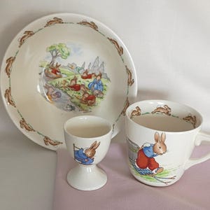 May include: A cream-colored ceramic dish, cup, and egg cup set. The dish has a border of brown rabbits and a central illustration of rabbits near a stream. The cup and egg cup feature rabbit illustrations. The set is designed for children.