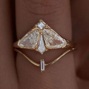 Może przedstawiać: Dwuczęściowy zestaw złotych pierścionków. Górny pierścionek ma unikalny design z trzema dużymi, przezroczystymi kamieniami w kształcie gruszki i małym kwadratowym kamieniem. Dolny pierścionek to zakrzywiona obrączka z małym, prostokątnym kamieniem.