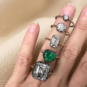 Op de afbeelding: Vijf ringen met gouden banden en verschillende edelsteen settings worden getoond. De edelstenen omvatten een hartvormige smaragd, een ronde diamant en rechthoekige en vierkante diamanten. De ringen zitten aan een vinger.