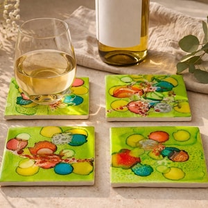 May include: Four square ceramic coasters with abstract designs in green, yellow, red, and blue. A wine glass with white wine sits on one coaster. A wine bottle is in the background.
