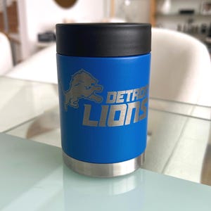 May include: A blue insulated can cooler with the Detroit Lions logo and text. The cooler has a black top and a silver base. The logo and text are in silver. The cooler is cylindrical and sits on a glass surface.