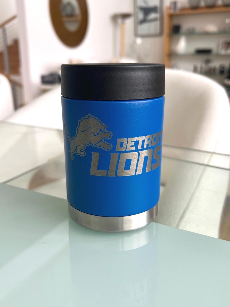 May include: A blue insulated can cooler with the Detroit Lions logo and text. The cooler has a black top and a silver base. The logo and text are in silver. The cooler is cylindrical and sits on a glass surface.