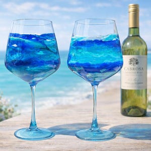 May include: Two clear wine glasses with blue liquid resembling ocean waves, next to a bottle of white wine. The wine bottle has a gold cap and a label that reads "CABROSA". The background shows a beach scene with the ocean and sky.