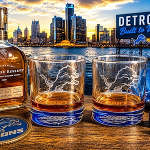 May include: A bottle of Woodford Reserve Kentucky Straight Bourbon Whiskey next to two clear glass tumblers, each etched with a lion logo. A blue and silver circular coaster and two grey stone whiskey cubes sit on a wooden surface.