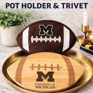 Op de afbeelding: Een bruine pannenlap en onderzetter set in de vorm van een voetbal. De pannenlap heeft witte strepen en een geel omlijnd blauw "M" logo. De onderzetter is van hout met het "M" logo en de tekst "University of Michigan".