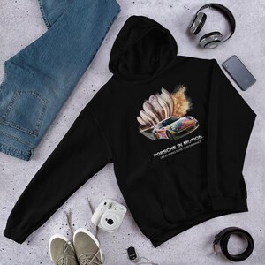 Könnte beinhalten: Ein schwarzer Kapuzenpullover mit einer Grafik eines bunten Sportwagens mit Bewegungsunschärfe-Effekt. Der Text unter dem Auto lautet PORSCHE IN MOTION. UNLEASHING PURE PERFORMANCE. Der Hoodie verfügt über lange Ärmel, eine Vordertasche und eine Kapuze.