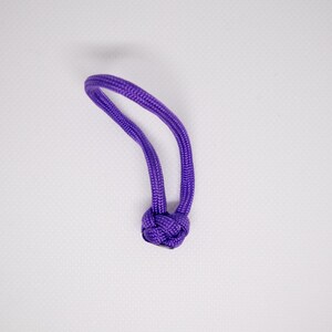 Puede incluir: Primer plano de un pequeño nudo de cuerda morada. La cuerda es de un tono morado vibrante y parece estar hecha de un material tejido apretado. El nudo está centrado en la imagen sobre un fondo blanco liso.