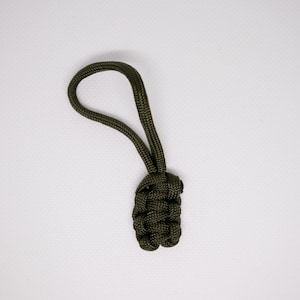Könnte beinhalten: Olivgrüner Paracord-Reißverschlussanhänger mit geflochtenem Knotenmuster. Der Anhänger hat eine Schlaufe zur einfachen Befestigung und einen dekorativen Knoten am Fuß. Geeignet für Taschen und Jacken.