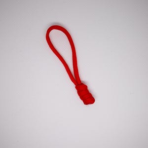 Könnte beinhalten: Ein leuchtend roter Reißverschluss-Anhänger mit einer Schlaufe oben und einer zylindrischen Basis. Der Anhänger besteht aus einem gewebten Material und befindet sich vor einem weißen Hintergrund. Dieser Artikel wird wahrscheinlich für Kleidung oder Taschen verwendet.
