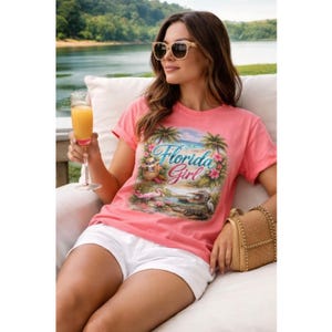 May include: A coral-colored t-shirt with the words "Florida Girl" and a tropical scene featuring palm trees, a flamingo, and a lake. The shirt is paired with white shorts and sunglasses.