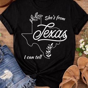 May include: Black t-shirt with a white graphic of the state of Texas and the words "she's from Texas, I can tell." The shirt is paired with denim shorts, sandals, and sunglasses on a wooden surface.