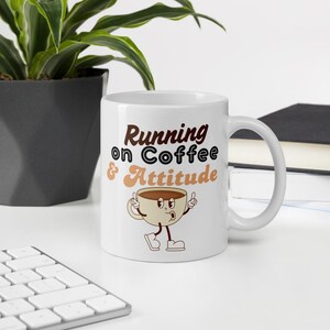 May include: White ceramic coffee mug with the text "Running on Coffee & Attitude" in brown and tan. The mug features a cartoon coffee cup character with arms and legs, giving a thumbs-up gesture.