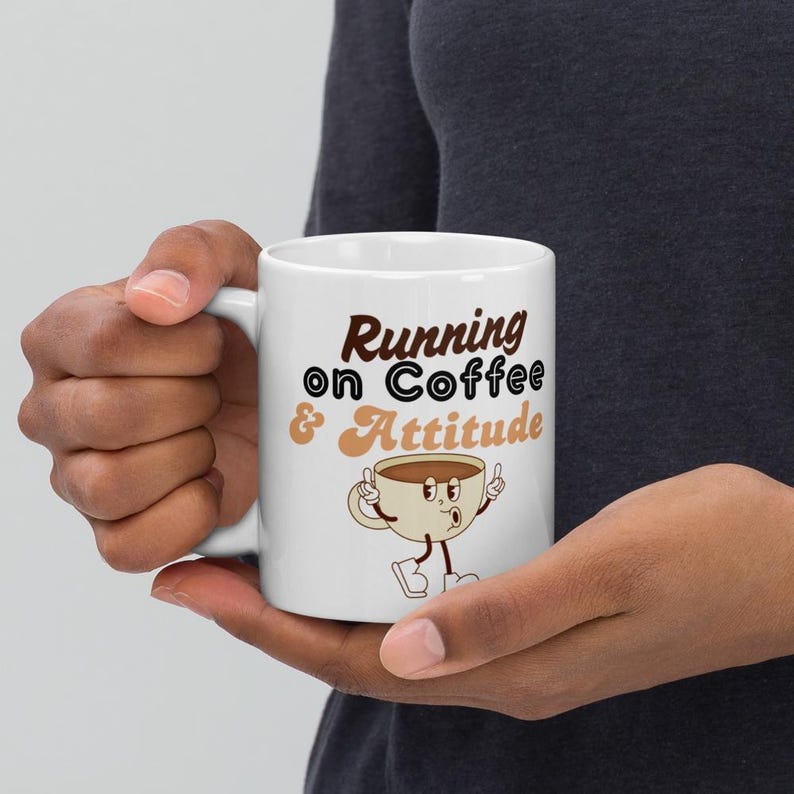 May include: White ceramic coffee mug with the text "Running on Coffee & Attitude" in brown and tan. The mug features a cartoon coffee cup character with arms and legs. The mug is held by a person.