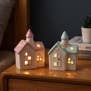 Puede incluir: Dos portavelas de cerámica con forma de casa. Uno es rosa y el otro azul, ambos con recortes de estrella y ventanas iluminadas. Las casas están sobre una superficie de madera, creando un ambiente cálido.