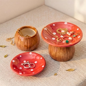 Puede incluir: Tres joyeros de cerámica con forma de hongo. El más grande es un hongo rojo con lunares blancos, que contiene collares, anillos y pendientes dorados. Los otros dos son un plato rojo pequeño con anillos y una olla marrón con una cadena dorada.