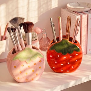 May include: Two ceramic strawberry-shaped holders, one pink and one red, are filled with makeup brushes and pens. The pink holder has yellow dots and green leaves, while the red one has white dots and green leaves. The holders sit on a white shelf.