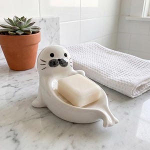 May include: A white ceramic soap dish shaped like a seal with a smooth finish, black painted eyes, and whiskers. The dish holds a rectangular bar of off-white soap. The seal figure has a rounded head and flippers extending to form the base.