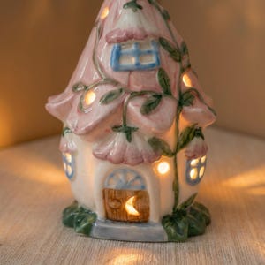 May include: A whimsical ceramic fairy house with a pink flower-shaped roof and white walls. The house features small blue-framed windows, a wooden door with a crescent moon, and glowing interior lights. Green vines and leaves decorate the exterior.