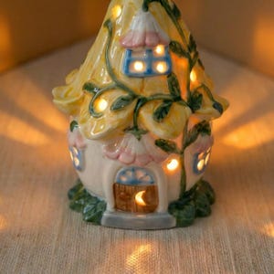 May include: A whimsical ceramic fairy house with a yellow roof, pink and blue accents, and green foliage. The house has glowing windows and a crescent moon above the door, creating a warm, inviting ambiance. A decorative accent piece.