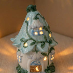 May include: A ceramic fairy house with a light blue roof and white walls. The house has multiple windows that glow with warm light. Green vines and leaves decorate the roof and walls, adding a whimsical touch. The door is a wooden style with a crescent moon.