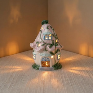 May include: A ceramic fairy house with a flower-shaped roof and windows. The house is painted in pastel colors with green vines and leaves. The windows and door glow with a warm light, creating a whimsical ambiance.