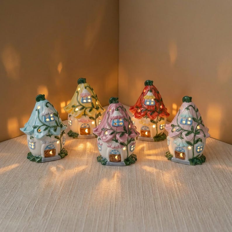 May include: A collection of five ceramic fairy houses in various colors, including blue, yellow, pink, and red. Each house features a floral design, lit windows, and a warm, inviting glow. The houses are arranged on a light-colored surface.